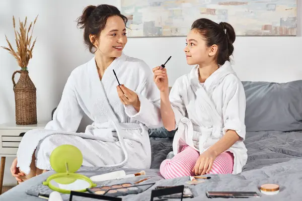 A brunette mother and daughter in white bath robes sitting comfortably on a bed, enjoying a peaceful moment together, holding mascara — Stock Photo