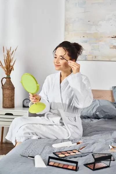 Eine Frau mit brünetten Haaren trägt Wimpern mit Mascara auf, während sie im weißen Bademantel auf einem Bett sitzt. — Stockfoto