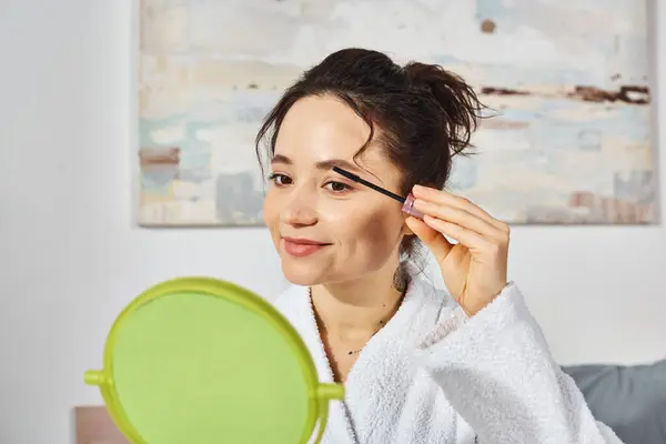 A woman with brunette hair is delicately applying mascara to her eyelashes while sitting on a bed in a white bath robe. — Stock Photo