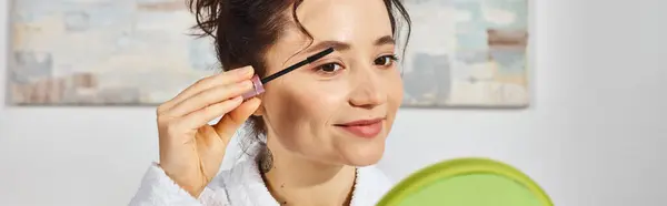 A brunette woman delicately applies mascara to her lashes while sitting on a bed in a white bath robe. — Stock Photo