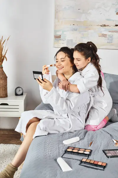 Una madre morena y su hija vestidas con batas blancas se sientan juntas en una cama acogedora, compartiendo un momento especial. - foto de stock
