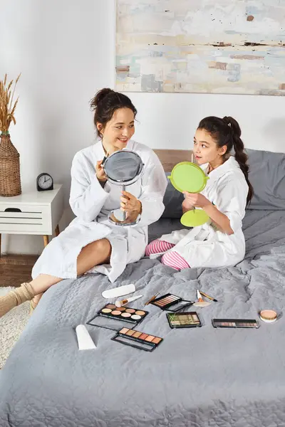 A brunette mother and her daughter, both dressed in white bath robes, sitting together on a cozy bed. — Stock Photo
