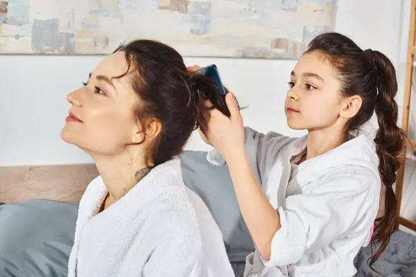 Uma mãe morena amorosamente penteia o cabelo de suas filhas jovens enquanto elas se sentam em uma cama com vestes de banho brancas. — Fotografia de Stock