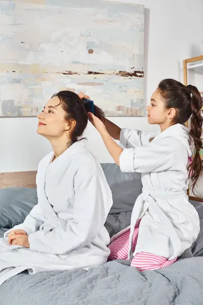 A brunette mother and daughter, both in white bath robes, sit peacefully on a cozy bed. — Stock Photo