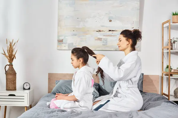 Morena mãe e filha em vestes de banho branco sentado na cama, compartilhando um momento amoroso como a mãe escova o cabelo das filhas. — Fotografia de Stock