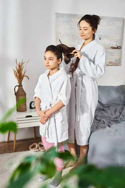 Una madre morena y su hija vestidas con batas de baño blancas de pie juntas en una habitación, compartiendo un momento especial. - foto de stock