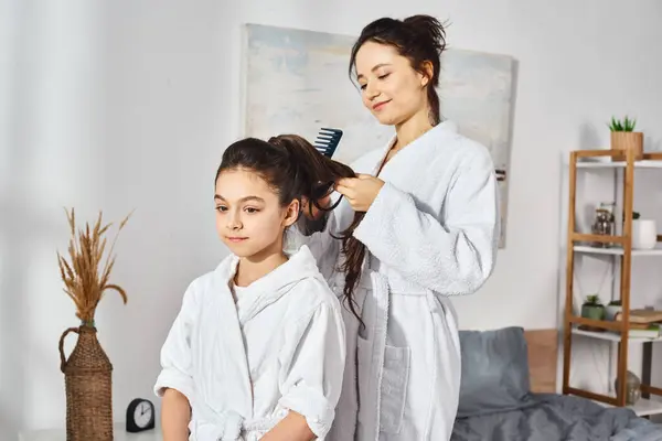 A brunette mother styled her daughters hair on a bed, both wearing white bath robes. — Stock Photo