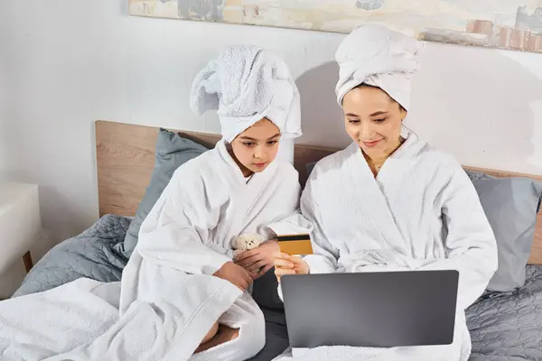 Two women, a brunette mother, and daughter, in white bath robes sitting on a bed, engaged with a laptop. — Stock Photo