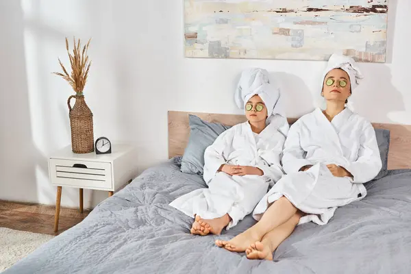 Брюнетка мать и дочь в белых халатах сидят на кровати, с полотенцами, обернутыми вокруг их голов. — стоковое фото