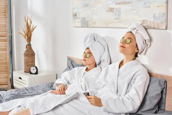 A brunette mother and daughter in white bath robes sitting comfortably on top of the bed, sharing a peaceful and intimate moment. — Stock Photo