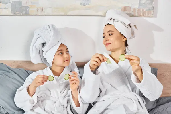 Brunette mother and daughter enjoy a peaceful moment while sitting on a bed in matching white robes, holding cucumbers, beauty — Stock Photo