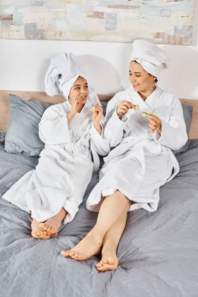 Una madre morena y una hija se sientan serenamente en una cama, cubiertas con túnicas blancas que fluyen, sosteniendo pepinos, belleza - foto de stock