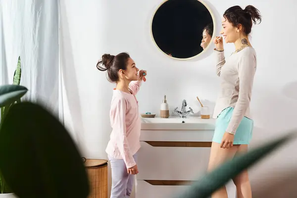 Une femme se brosse les dents devant un miroir tandis que sa fille regarde dans une salle de bain moderne. — Photo de stock
