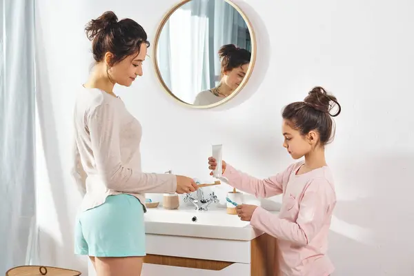 Una morena y su hija preadolescente están juntas frente a un lavabo en un baño moderno, dedicándose a rutinas de belleza e higiene. - foto de stock