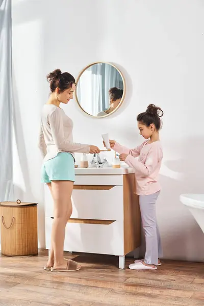 Una mujer morena y su hija preadolescente están en un baño moderno, dedicándose a su rutina de belleza e higiene junto al fregadero. - foto de stock