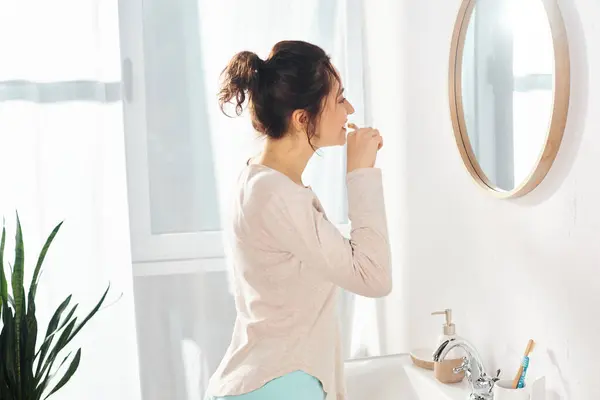 Una morena se cepilla los dientes delante de un espejo, mientras se encuentra en un baño moderno. - foto de stock
