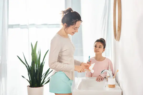 Una morena y su hija preadolescente están paradas una al lado de la otra en un moderno lavabo de baño, dedicadas a una rutina de belleza e higiene.. - foto de stock