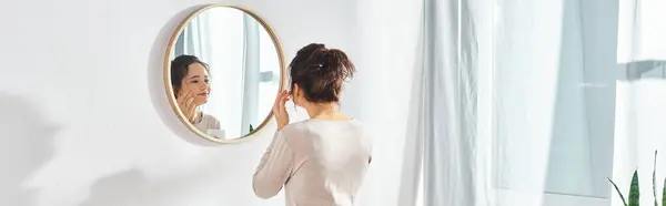 Una donna bruna si trova di fronte a uno specchio, applicando crema per il viso in un bagno moderno. — Foto stock