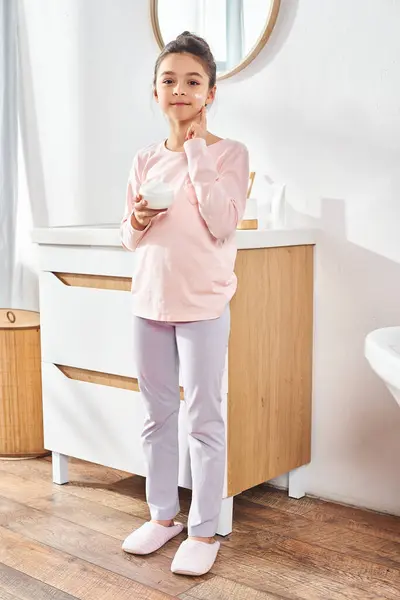 A little girl with brown hair stands in front of a sleek bathroom sink, exploring her beauty, applying cream — Stock Photo