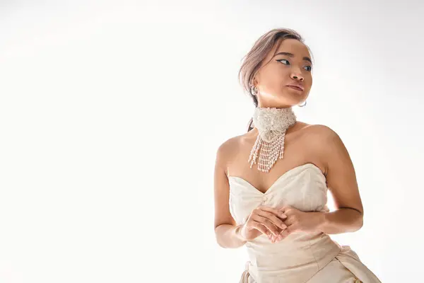 Elegante asiático joven mujer en blanco vestido restrained posando y mirando a lado en blanco fondo - foto de stock