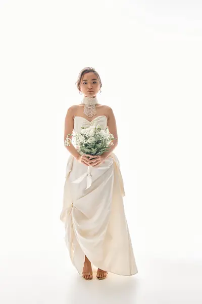 Витончена азіатська наречена в елегантній сукні, що тримає букет з білими квітами і дивиться на камеру — Stock Photo