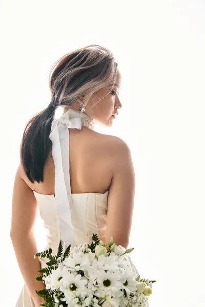 Verführerische junge Braut im weißen Kleid mit Blumenstrauß hinter ihrem Rücken auf hellem Hintergrund — Stockfoto