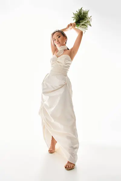 Encantadora mujer joven asiática en vestido blanco a punto de lanzar ramo de flores en fondo claro - foto de stock