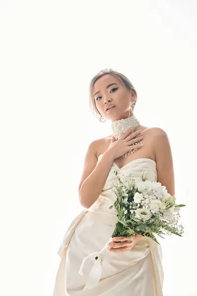 Привлекательная азиатская женщина прикоснуться к белому ожерелью и протянуть букет цветов на светлом фоне — стоковое фото