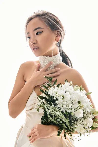 Encantadora mujer asiática tocando a collar blanco y mirando a un lado con ramo de flores - foto de stock