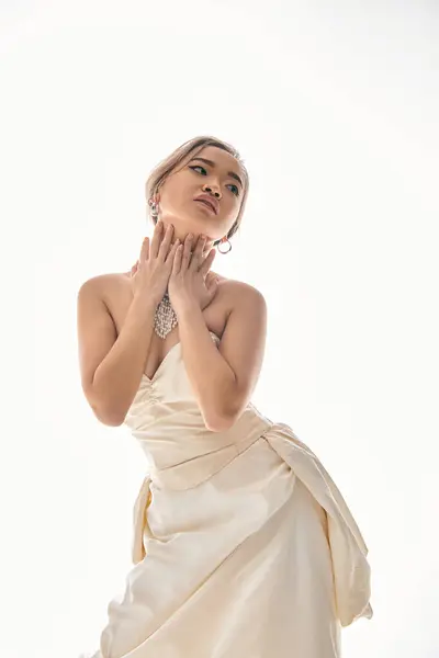 Alluring asian bride in elegant dress look to side and hug neck with hands on light background — Stock Photo