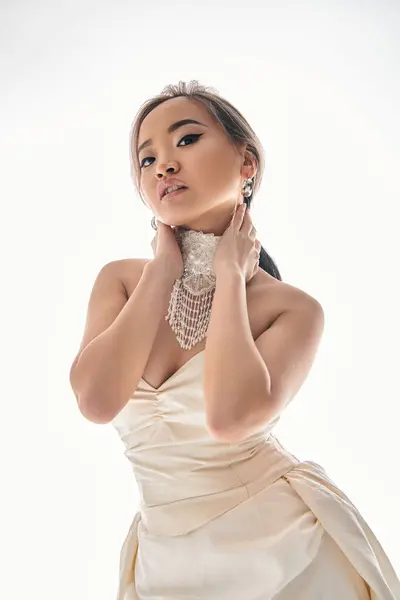 Elegante asiático joven mujer en blanco vestido look a cámara y abrazo cuello con manos sobre fondo claro - foto de stock
