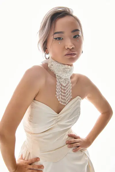 Selbstbewusste asiatische junge Braut in eleganter Halskette und Kleid posiert auf weißem Hintergrund — Stockfoto