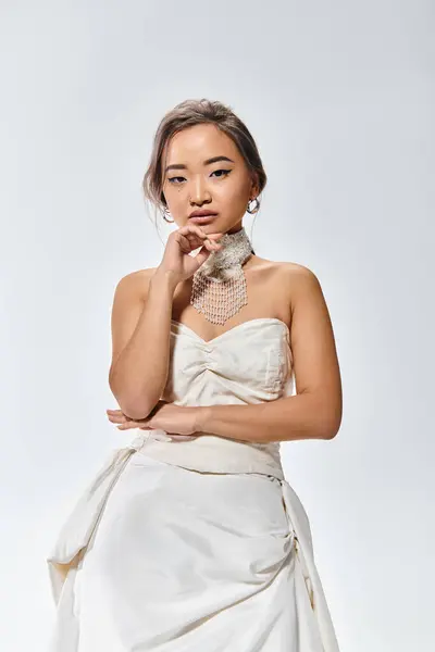 Elegante mujer joven asiática en vestido elegante con expresión reflexiva en la cara sobre fondo claro - foto de stock