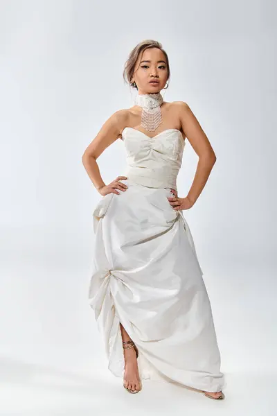 Atractivo asiático joven novia en blanco elegante vestido confiado posando sobre fondo claro - foto de stock