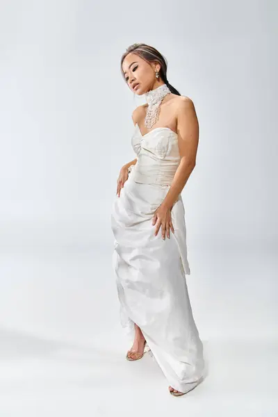 Bonita mujer asiática en blanco elegante vestido posando y mirando hacia abajo sobre fondo claro - foto de stock