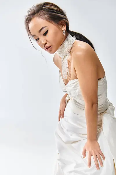 Encantadora chica asiática en elegante vestido blanco inclinado hacia adelante y mirando hacia abajo sobre fondo claro - foto de stock