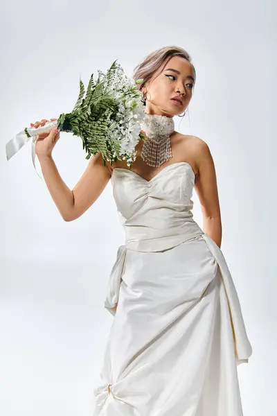 Atractiva novia asiática en traje elegante blanco posando con flores ramo sobre fondo claro - foto de stock