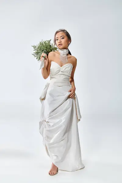 Belle femme asiatique en tenue de grâce blanche posant avec bouquet de fleurs sur fond clair — Photo de stock