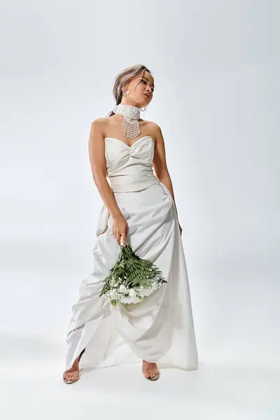 Atractiva joven novia en traje elegante blanco girando cabeza y posando con ramo de flores - foto de stock