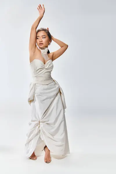 Alluring asian bride in white grace dress posing with hands and looking to side on light background — Stock Photo
