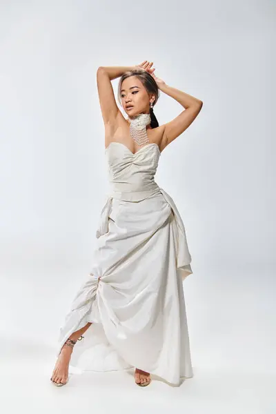 Elegante asiático joven mujer en novia traje buscando abajo y poniendo manos detrás de la cabeza - foto de stock