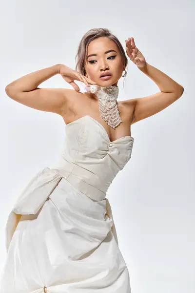 Elegante asiático mujer en blanco gracia vestido posando con las manos cerca de cara y mirando hacia abajo - foto de stock
