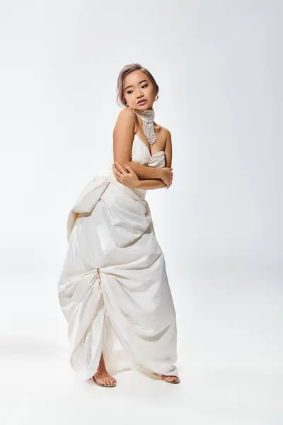 Graceful asian young woman in white elegant dress hug herself and look to down on light background — Stock Photo