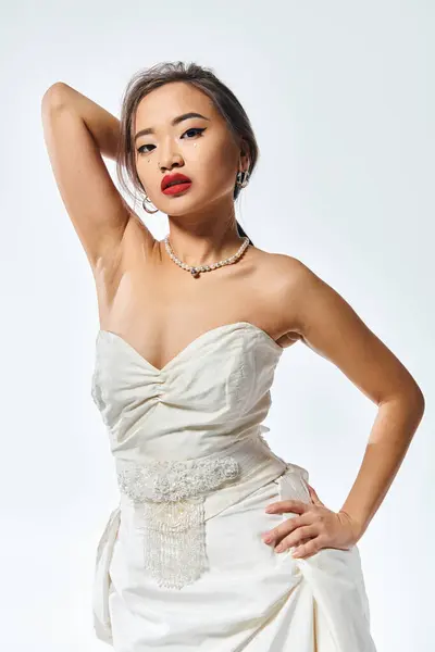 Gracieuse asiatique femme en nuptiale tenue avec rouge lèvres mettre la main derrière la tête sur fond blanc — Photo de stock