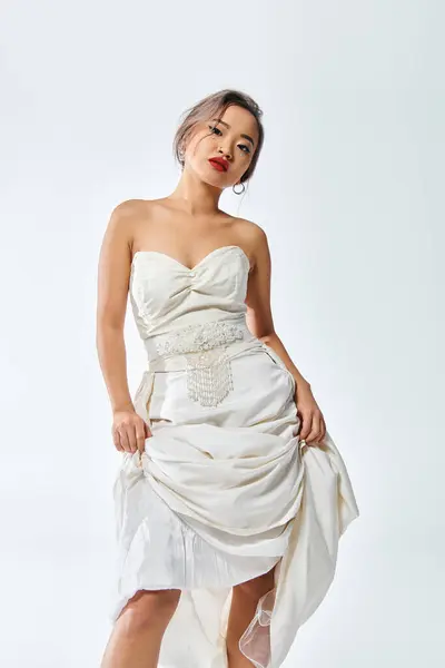 Elegante mujer asiática en traje blanco elegante con labios rojos cabeza inclinada y levantó su vestido - foto de stock