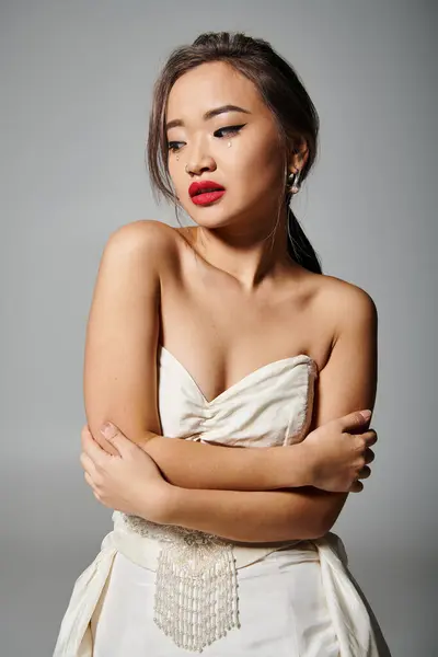 Seductora chica joven asiática con collar de perlas y lápiz labial rojo abrazarse sobre fondo gris - foto de stock