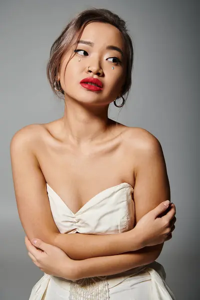 Очаровательная азиатская молодая женщина с красной помадой обнять себя и смотреть в сторону на сером фоне — стоковое фото