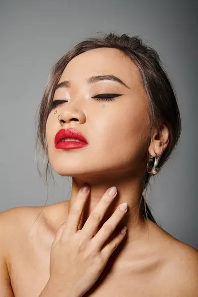 Elegante asiático chica en su 20s con cerrado ojos y rojo lápiz labial abrazo a cuello en gris fondo - foto de stock