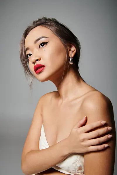 Porträt einer attraktiven asiatischen Frau, die sich an der Schulter festhält und nach oben schaut auf grauem Hintergrund — Stockfoto