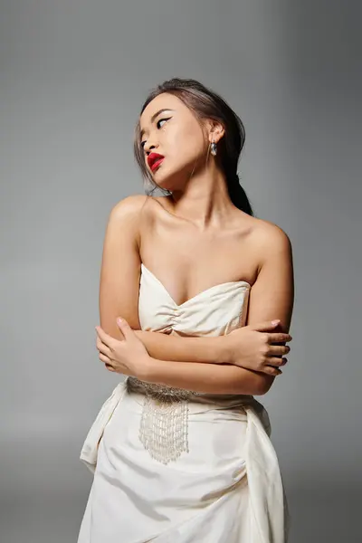 Seductora mujer joven asiática en vestido blanco abrazarse y mirar a un lado sobre fondo gris - foto de stock